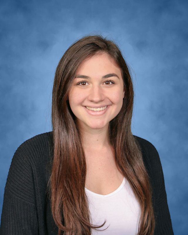 A young woman with long brown hair, wearing a white top and dark sweater, smiles in front of a blue studio background.