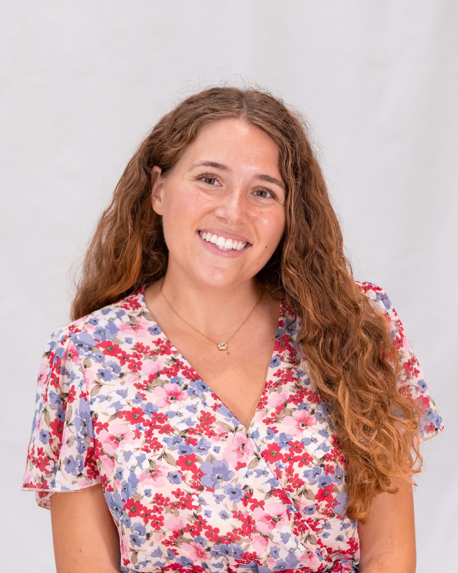A woman with long curly hair wearing a floral dress smiles at the camera against a plain light background.