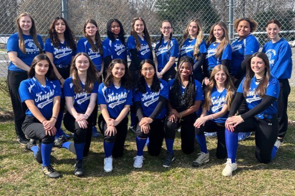 A group of sixteen young women in blue and black softball uniforms pose for a team photo outdoors in front of a chain-link fence.