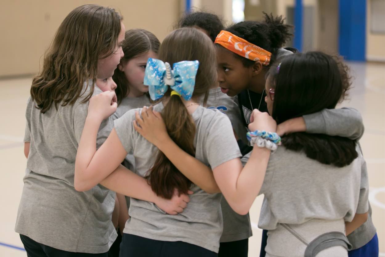 A group of girls in sportswear stand in a huddle, arms around each other, having a discussion in a gymnasium.