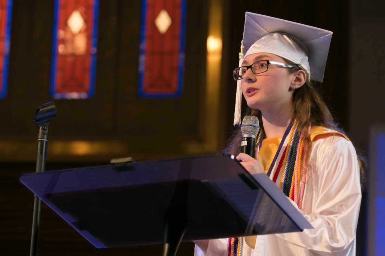 A young graduate in cap and gown speaks at a podium with a microphone in front of stained glass windows.