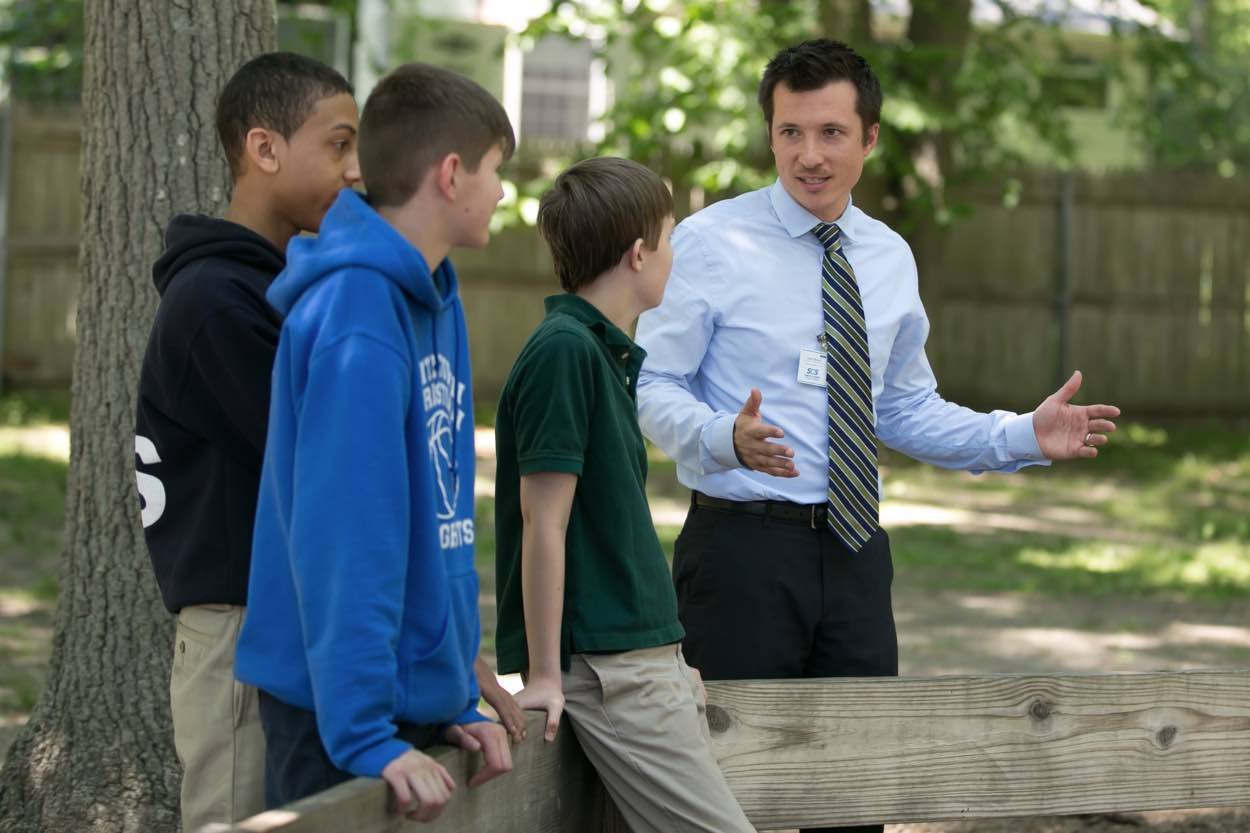 A man in a dress shirt and tie talks to three boys wearing school uniforms outdoors near a wooden fence and trees.