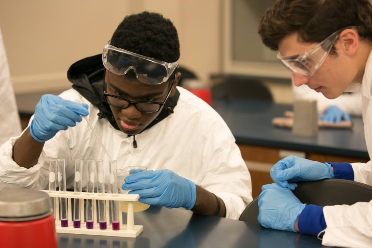 Two students wearing lab coats, safety goggles, and gloves conduct an experiment with test tubes and a petri dish in a science laboratory.