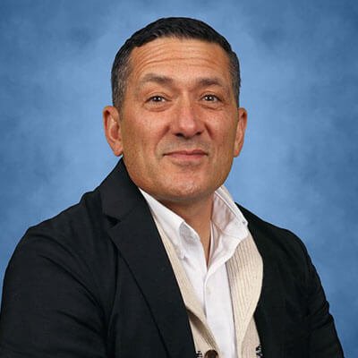 A man with short dark hair wearing a white shirt and black jacket poses in front of a blue studio background.