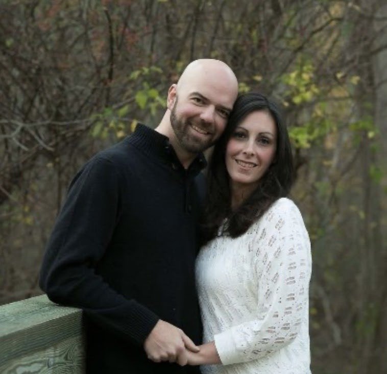 A man and woman stand close together outdoors by a wooden railing, smiling at the camera with trees in the background.