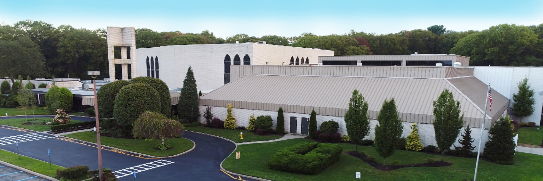 A large, modern building with white walls, pointed windows, and neatly landscaped bushes and trees, adjacent to a parking lot with marked spaces.