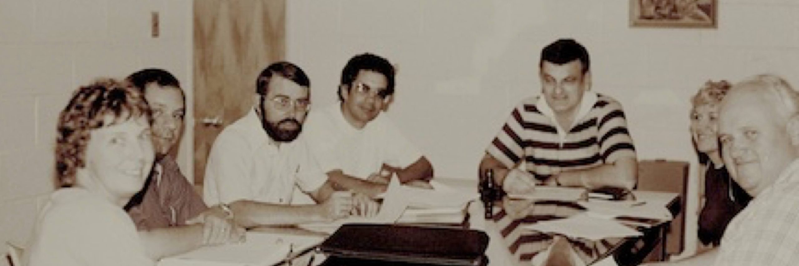 Seven people sit around a table in a meeting room, engaged in discussion with papers and folders in front of them.