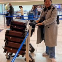 A person in a gray coat and jeans stands in an airport, pushing a luggage cart stacked with four suitcases. Other travelers are seen in the background.