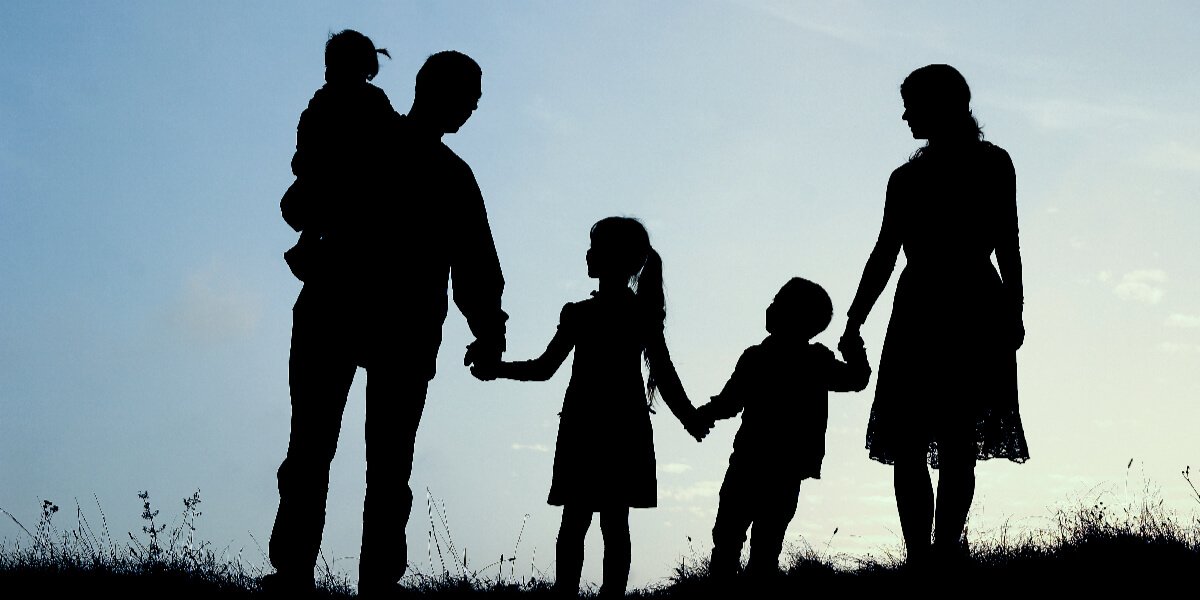 Silhouette of two adults and three children holding hands outdoors against a blue sky.