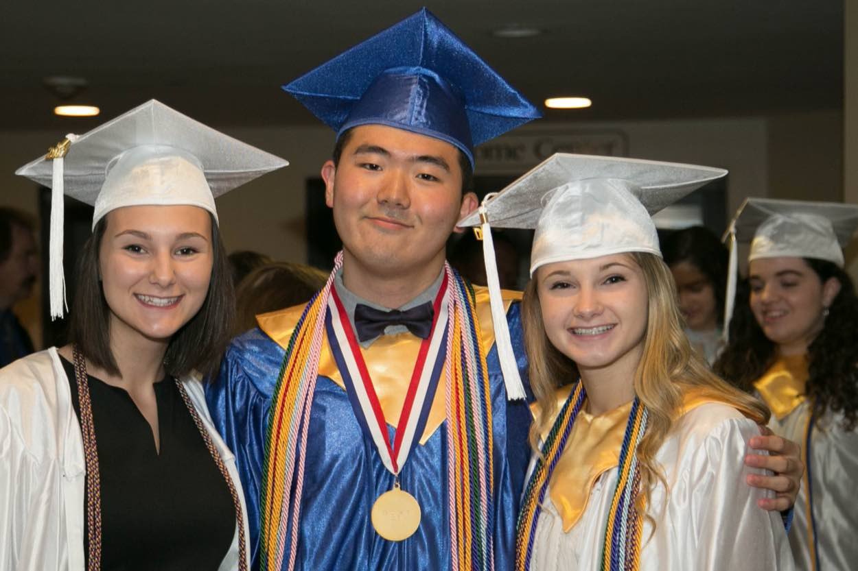 Three students in graduation caps and gowns pose together, smiling. The student in the center wears a blue cap and gown with multiple honor cords and medals.