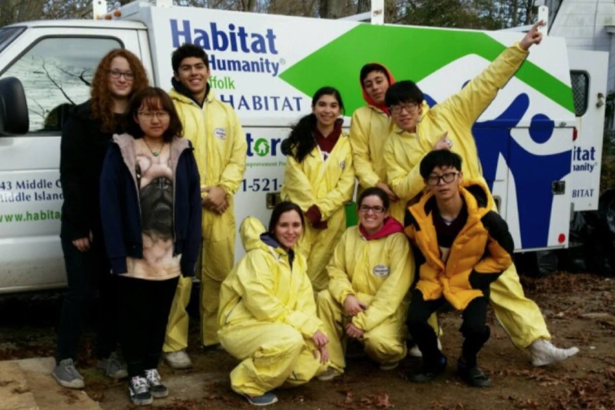 A group of eight young people, some in yellow coveralls, pose in front of a Habitat for Humanity van outdoors.