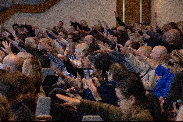 A large group of people seated in an auditorium raise their right arms forward in unison.