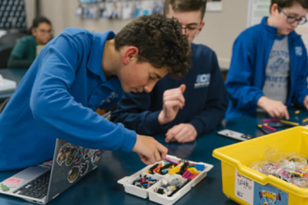 A student in a blue shirt selects pieces from a plastic container of robotics parts while others work at the same table.