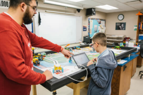 An adult points at a robotics game board while a student works on a tablet in a classroom setting.