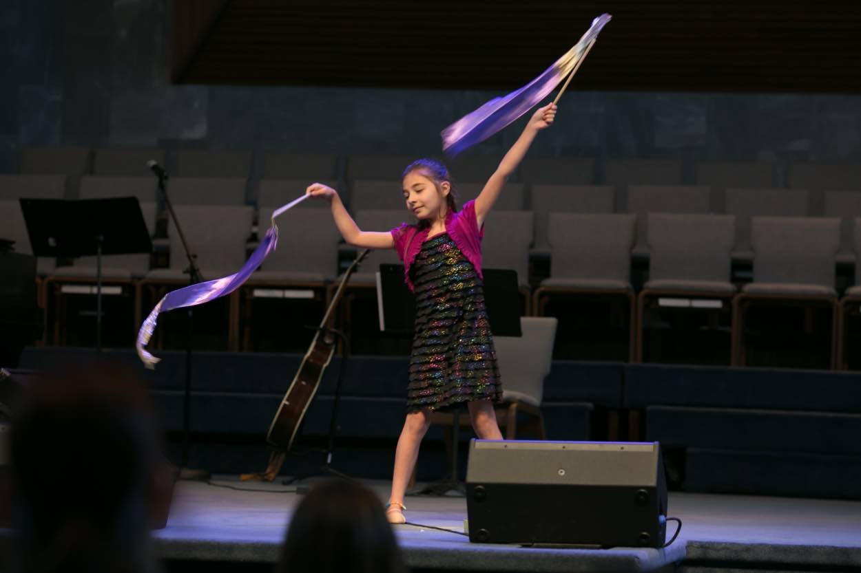 A young girl in a dress performs a ribbon dance on stage, with chairs, a guitar, and a microphone visible in the background.
