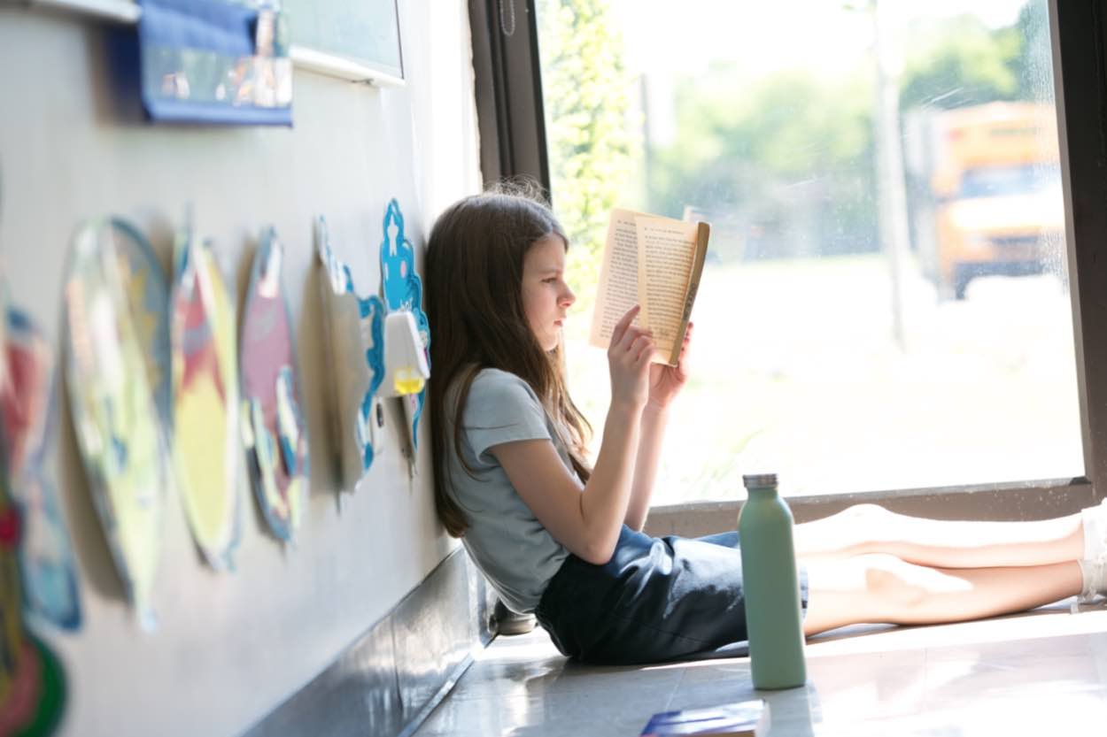 A young girl sits on the floor by a window, reading a book with a water bottle placed nearby. Sunlight streams in through the glass.