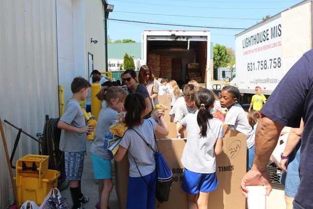 Children and adults gather around boxes, sorting items near a truck outside a building labeled "Lighthouse Mission.